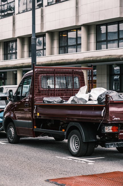 A small burgundy flatbed truck parked on a city street with its cargo area filled with various black and white plastic bags and loose waste materials. The truck features a high metal mesh barrier behi