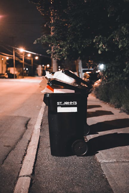 A large blue plastic rubbish bin with a closed lid positioned on a grassy outdoor area, adjacent to a red wheeled waste container with black ventilation holes on its side. The blue bin features a yell